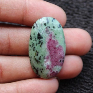 May include: Oval-shaped green and pink gemstone with black flecks. The gemstone is smooth and polished.