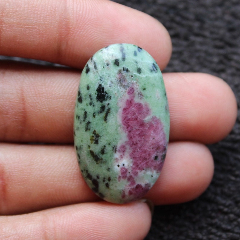 May include: Oval-shaped green and pink gemstone with black flecks. The gemstone is smooth and polished.