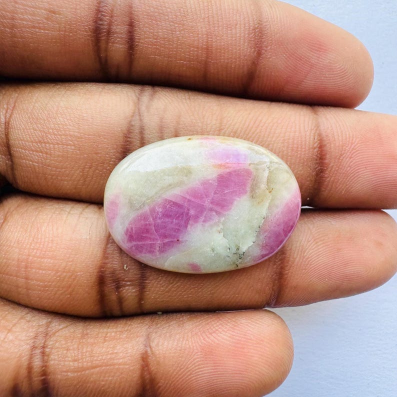 Pu&ograve; includere: Gemma ovale con superficie lucida. La pietra presenta un mix di tonalit&agrave; rosa e bianche, con striature rosa che attraversano la base bianca. La gemma &egrave; tenuta in una mano.