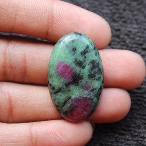 May include: Oval-shaped green gemstone with black and pink inclusions. The gemstone is set in a cabochon cut.