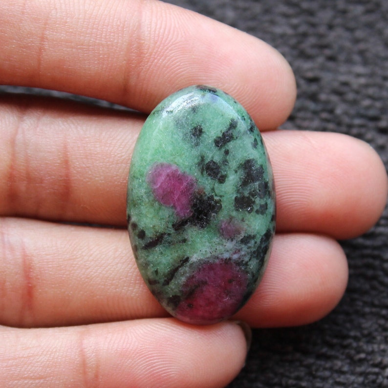May include: Oval-shaped green gemstone with black and pink inclusions. The gemstone is set in a cabochon cut.