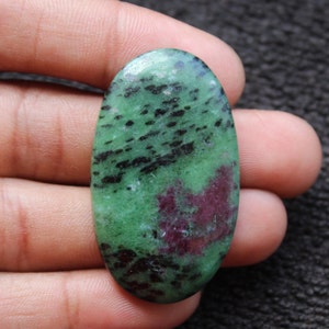 May include: Oval-shaped green gemstone cabochon with black and pink inclusions. The stone has a smooth, polished surface.
