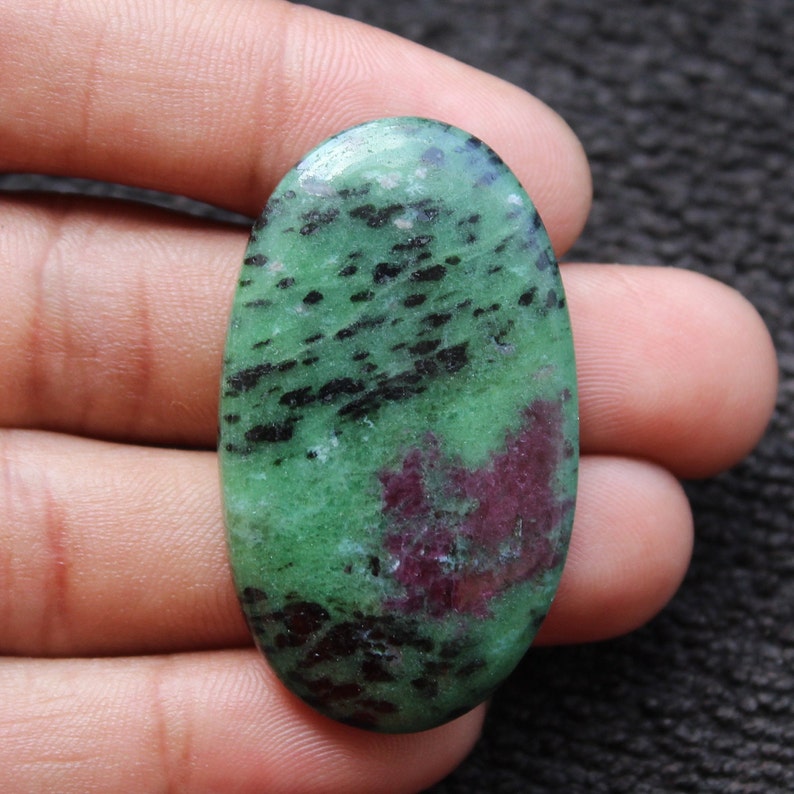 May include: Oval-shaped green gemstone cabochon with black and pink inclusions. The stone has a smooth, polished surface.