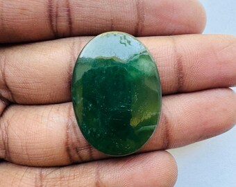 May include: An oval-shaped, polished gemstone with a deep green hue, featuring lighter green and yellow inclusions. The stone is smooth and reflective, showcasing natural variations in color and texture. The gemstone is held in a hand.