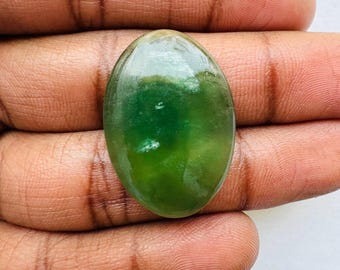 May include: An oval-shaped, polished gemstone with a gradient of green hues. The stone has a smooth, glossy surface, and the color transitions from a darker green to a lighter, almost translucent shade. The gemstone is held in a hand.