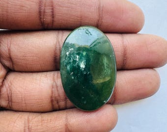 May include: An oval-shaped, polished gemstone with a deep green hue and lighter green inclusions. The stone is held in a hand, showcasing its smooth surface and natural variations. This gemstone could be used for jewelry or other crafts.
