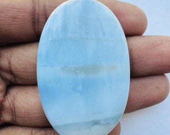 May include: An oval-shaped, polished gemstone with a light blue and white color gradient. The stone has a smooth surface with subtle banding, creating a layered effect. The gemstone is held in a hand.