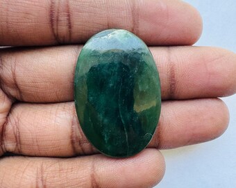 May include: An oval-shaped, polished gemstone with a deep green hue. The stone has a smooth surface with variations in color, ranging from dark to lighter shades of green. The gemstone is held in a hand, showcasing its size and shape.