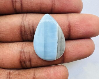 May include: A teardrop-shaped gemstone with a light blue and gray color scheme. The stone is divided into two sections, with the left side a solid light blue and the right side a mix of gray and blue. The stone is held in a hand.