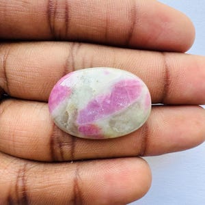 Pu&ograve; includere: Gemma ovale con superficie lucida. La pietra presenta un mix di tonalit&agrave; rosa e bianche, con il rosa che forma motivi irregolari. La gemma &egrave; tenuta in una mano, mostrando le sue dimensioni e la sua forma.