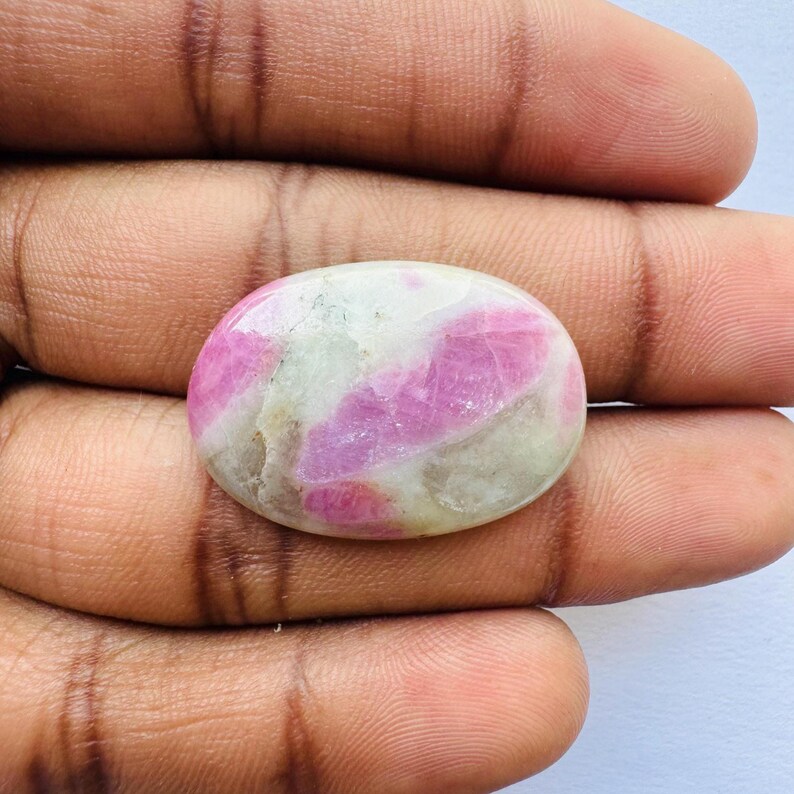 Pu&ograve; includere: Gemma ovale con superficie lucida. La pietra presenta un mix di tonalit&agrave; rosa e bianche, con il rosa che forma motivi irregolari. La gemma &egrave; tenuta in una mano, mostrando le sue dimensioni e la sua forma.