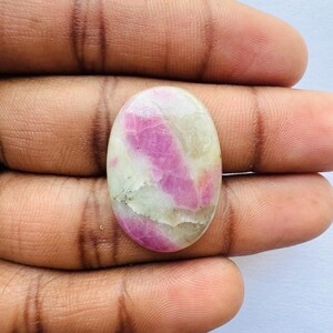 Pu&ograve; includere: Gemma ovale con una miscela di colori rosa e crema. La pietra lucida mostra venature rosa su una base color crema. La gemma &egrave; tenuta in mano, evidenziando le sue caratteristiche naturali.