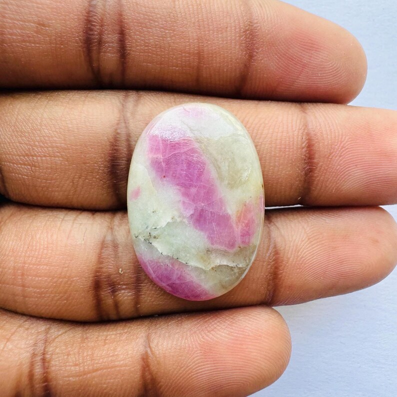 Pu&ograve; includere: Gemma ovale con una miscela di colori rosa e crema. La pietra lucida mostra venature rosa su una base color crema. La gemma &egrave; tenuta in mano, evidenziando le sue caratteristiche naturali.