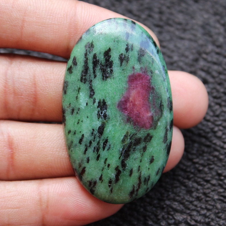 May include: Oval-shaped green gemstone with black and pink inclusions. The stone has a smooth, polished surface.