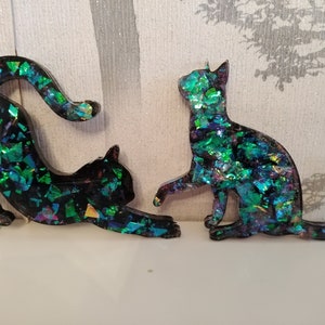 Può includere: Due statuette di gatti neri con glitter iridescenti verdi e blu incorporati nella resina. Un gatto si allunga, l'altro è seduto. Lo sfondo è grigio chiaro e bianco.