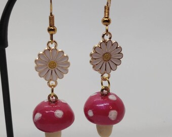 May include: A pair of dangle earrings featuring a daisy and mushroom design. The earrings have gold-colored hooks and connectors. The daisies are white with gold centers, and the mushrooms are red with white spots and beige stems.
