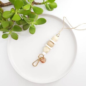 May include: A beige and white beaded keychain with a gold charm. The keychain is on a white plate with a green plant in the background.