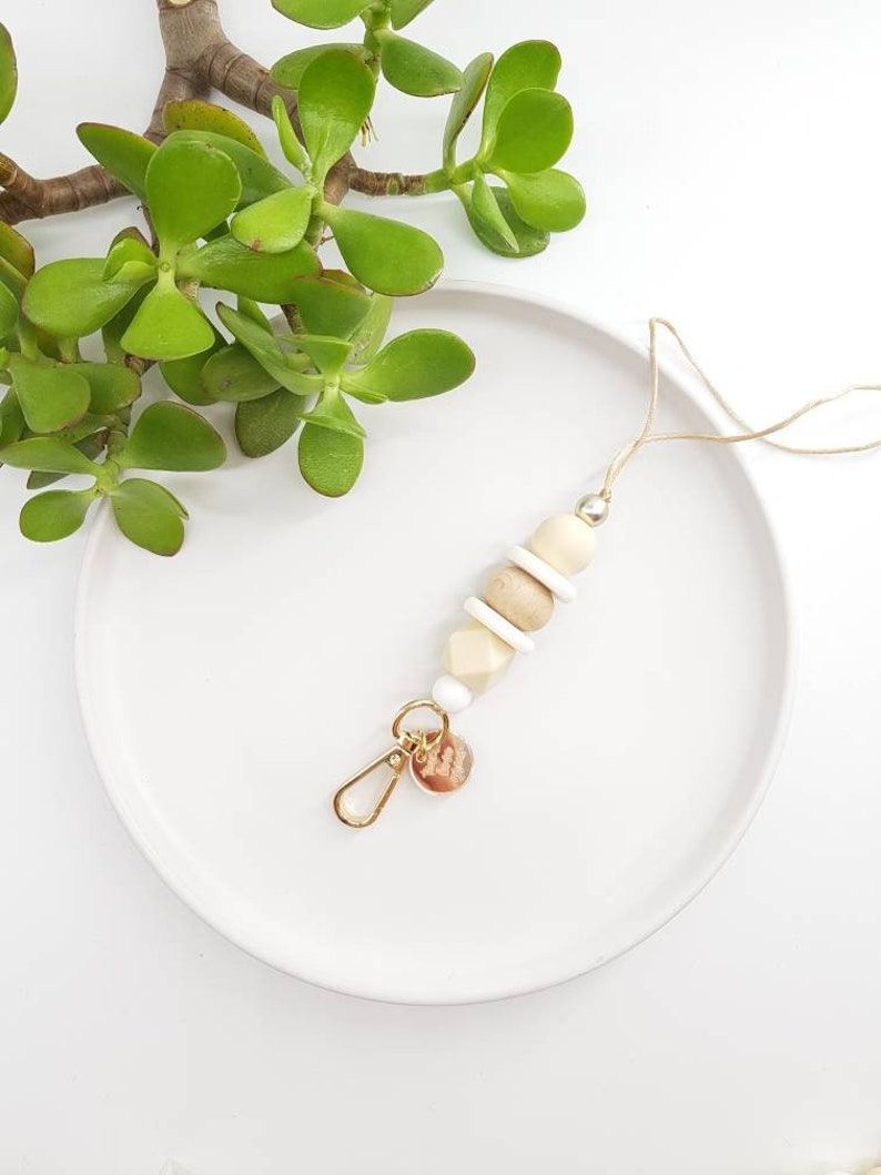 May include: A beige and white beaded keychain with a gold charm. The keychain is on a white plate with a green plant in the background.