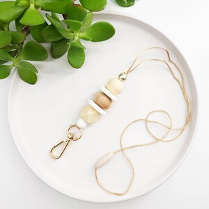 May include: A beige and white beaded lanyard with a gold clasp and a wooden bead accent. The lanyard is on a white plate with a green plant in the background.