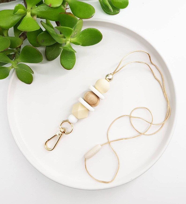 May include: A beige and white beaded lanyard with a gold clasp and a wooden bead accent. The lanyard is on a white plate with a green plant in the background.