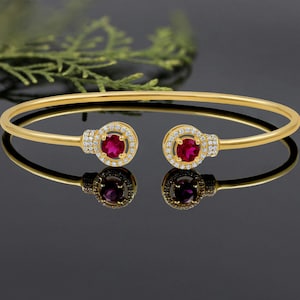 Può includere: Un bracciale rigido dorato con due elementi circolari con rubini e diamanti. I rubini sono di un rosso intenso, circondati da un alone di piccoli diamanti trasparenti. Il bracciale è esposto su una superficie nera riflettente.