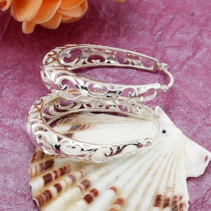 May include: A pair of silver hoop earrings with an open filigree design. The earrings are displayed on a seashell against a textured purple background, with peach-colored flowers in the background.
