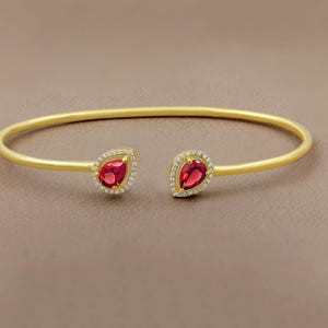 Può includere: Un bracciale aperto color oro con due gemme rosse a forma di goccia. Ogni gemma è circondata da un alone di piccoli cristalli trasparenti. Il bracciale presenta un design semplice ed elegante.