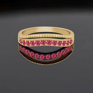 Peut inclure: Une bague dorée ornée d'une rangée de petits pierres précieuses roses vives. La bague a un design à double bande, avec une bande supérieure texturée et une bande inférieure avec les pierres précieuses. La bague se reflète sur une surface noire.