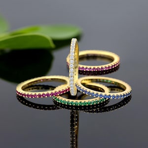 Peut inclure: Quatre bagues en or avec une rangée de petites pierres précieuses. Les bagues sont empilées les unes sur les autres. La bague du haut a des pierres précieuses claires, la deuxième bague a des pierres précieuses rouges, la troisième bague a des pierres précieuses bleues et la bague du bas a des pierres précieuses vertes.
