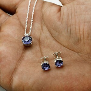 Puede incluir: Un collar de plata con un colgante de piedra preciosa p&uacute;rpura y pendientes a juego. El collar tiene una cadena delicada y el colgante es una piedra preciosa redonda y facetada.