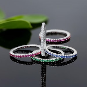 May include: Four silver stacking rings with a single row of colorful gemstones. The rings are stacked with a white ring on top, followed by a red ring, a blue ring, and a green ring.