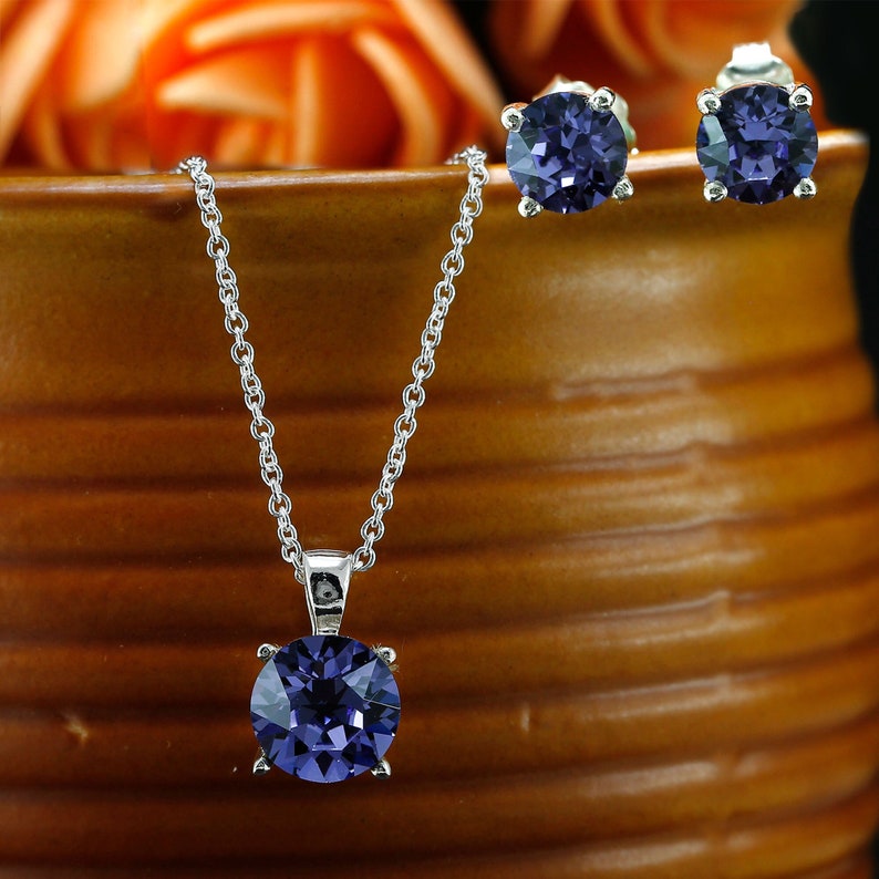 Puede incluir: Un collar de plata con un colgante redondo de piedra preciosa de color p&uacute;rpura oscuro y pendientes a juego. El collar tiene una cadena delicada y los pendientes son de estilo de bot&oacute;n.