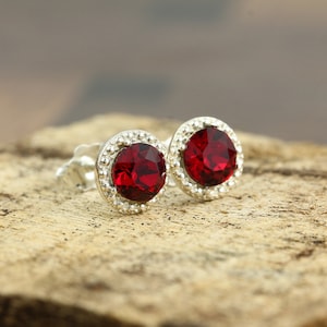 May include: A pair of silver stud earrings with a red crystal center and a silver halo around the crystal.