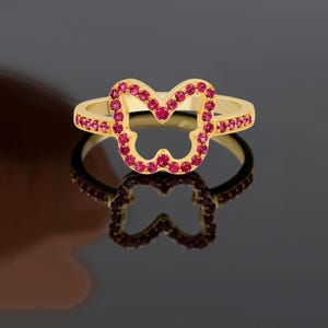 May include: A gold ring with a butterfly-shaped design, studded with small, round, red gemstones. The ring's band also features small red gemstones. The ring is shown on a reflective surface, creating a mirrored effect.