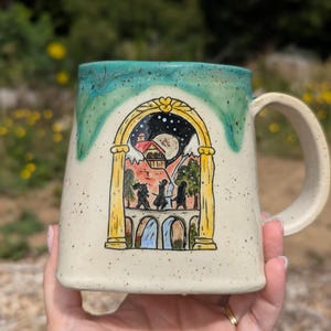 May include: A ceramic mug with a speckled cream base and a teal drip glaze at the rim. The front features a hand-painted illustration of three figures walking across a stone bridge toward a building, framed by a yellow archway under a night sky.