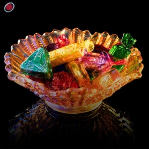 May include: A clear glass bowl with a scalloped rim filled with colorful wrapped chocolates. The bowl is sitting on a black surface.