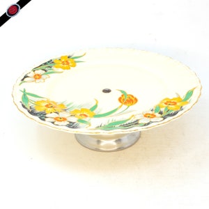 May include: A white cake stand with a silver base. The stand is decorated with a floral pattern of yellow, orange, and green flowers.