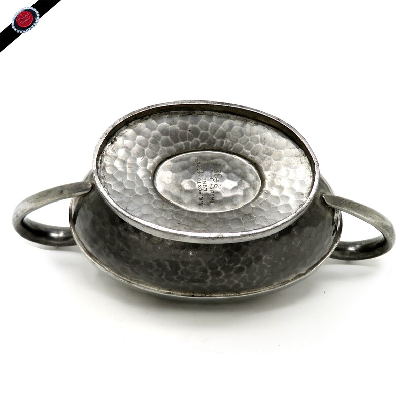 May include: A silver-toned, hammered metal sugar bowl with two handles. The bowl has an oval base and a textured surface. The underside of the bowl features stamped text, including "A.E. POSTON & CO. LONDON ENGLISH PRO 243".