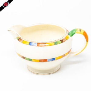 May include: A white ceramic creamer with a colorful geometric pattern around the rim. The creamer has a rounded body and a small handle.