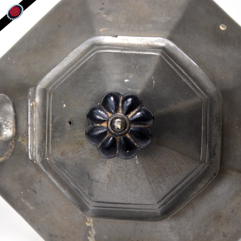 May include: A silver teapot with a black, flower-shaped knob on the lid. The teapot has an octagonal shape and a spout on the side.