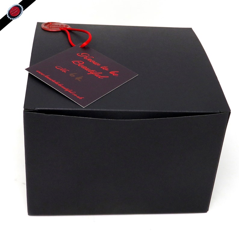 May include: A black, square gift box with a red tag attached. The tag reads "Know to be Beautiful" in red script, with a website address below. A red wax seal is also attached to the box.