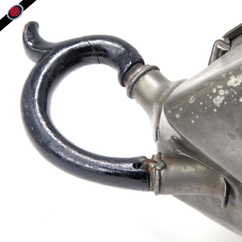 May include: A pewter teapot with a black wooden handle. The teapot has a unique, angular shape and a spout with a decorative design.