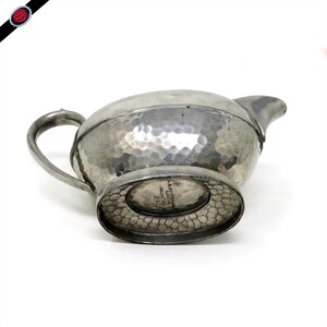 May include: A silver-colored, hammered metal creamer with a spout and handle. The creamer has a textured surface and a decorative base. The bottom of the creamer has text that reads "A.E.P. STREETER ENGLISH PEWTER LONDON 2+5".