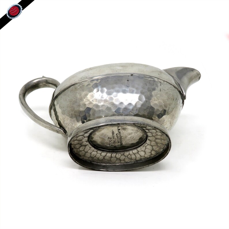 May include: A silver-colored, hammered metal creamer with a spout and handle. The creamer has a textured surface and a decorative base. The bottom of the creamer has text that reads "A.E.P. STREETER ENGLISH PEWTER LONDON 2+5".