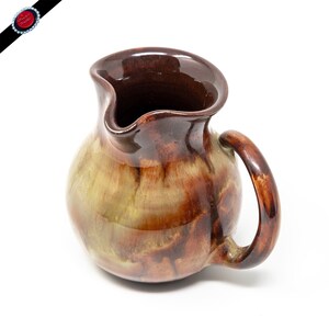 May include: A brown and yellow ceramic pitcher with a rounded body and a handle. The pitcher has a glazed finish with a mottled pattern.