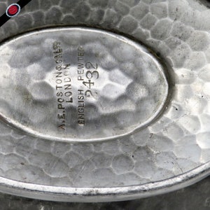 May include: Close-up of a vintage English pewter dish with a hammered texture. The oval center features the text "A.E. POSTON & CO. LTD LONDON ENGLISH PEWTER 2432". The dish has a slightly raised rim and a dull silver color.