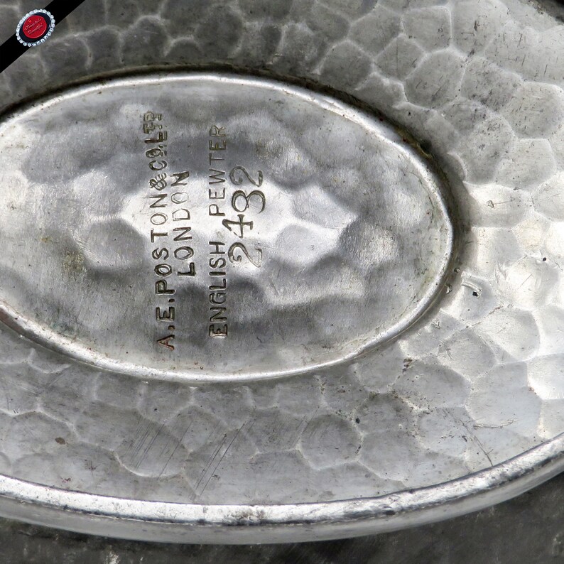 May include: Close-up of a vintage English pewter dish with a hammered texture. The oval center features the text "A.E. POSTON & CO. LTD LONDON ENGLISH PEWTER 2432". The dish has a slightly raised rim and a dull silver color.