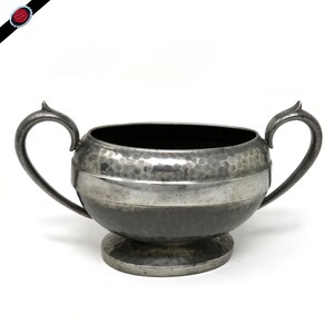 May include: A silver-toned, hammered metal sugar bowl with two curved handles. The bowl has a wide, horizontal band of polished metal and sits on a small, round base. The interior is dark, contrasting with the exterior's metallic sheen.