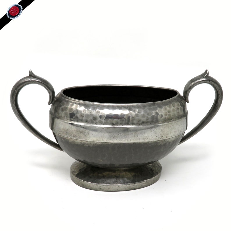 May include: A silver-toned, hammered metal sugar bowl with two curved handles. The bowl has a wide, horizontal band of polished metal and sits on a small, round base. The interior is dark, contrasting with the exterior's metallic sheen.