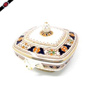 May include: A white porcelain serving dish with a lid. The dish has a square shape with rounded corners and features a decorative design in blue, orange, green, and gold. The lid has a finial in the shape of a stylized flower.
